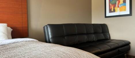 A hotel room with a bed, a black leather sofa, a wooden headboard, and a framed artwork on the wall.