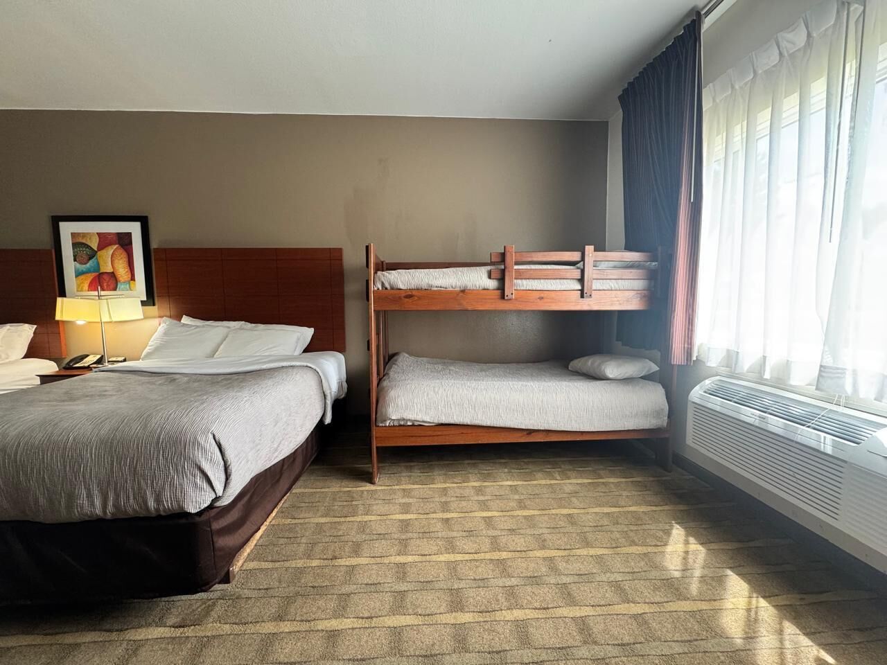 A hotel room with two beds, a bedside table with a lamp, a painting on the wall, and a window with curtains.