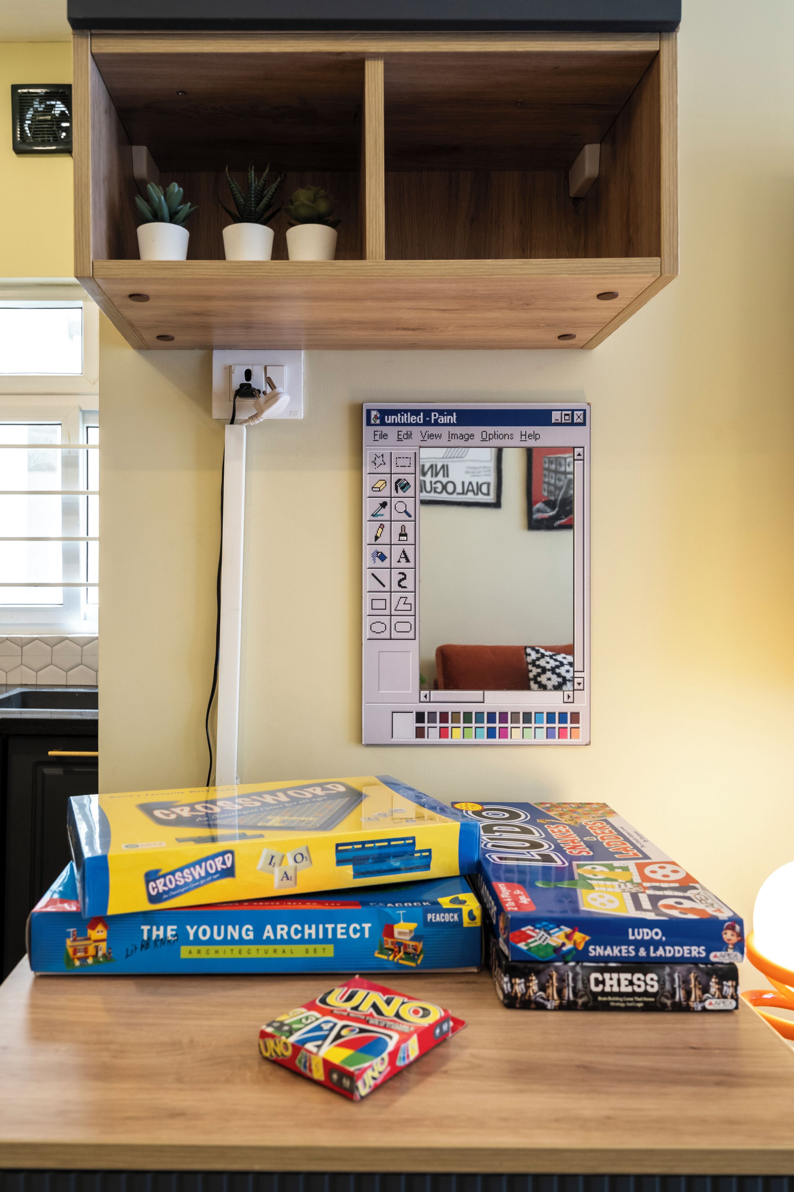 Un bureau avec des jeux de société, un écran d’ordinateur et une étagère en bois ornée de plantes.