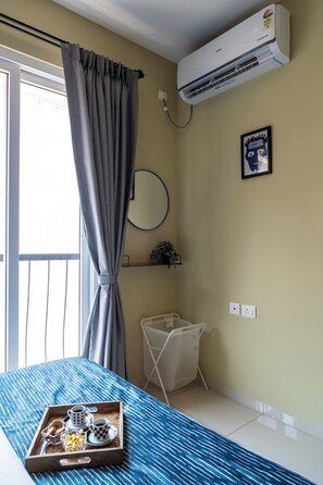Une chambre d’hôtel avec un lit, un plateau contenant des articles pour le petit-déjeuner, un climatiseur, une fenêtre avec des rideaux, un miroir fixé au mur et une petite étagère avec une plante.