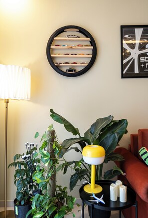 Une pièce avec une étagère murale ronde où sont exposées des voitures miniatures, une lampe jaune, une plante en pot et un tableau encadré.