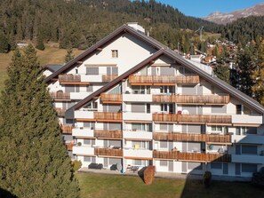 Ein mehrgeschossiges Gebäude mit Balkonen und einer flachen Dachterrasse, vor einer gebirgigen Kulisse.