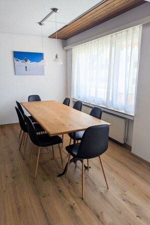 Uma sala de conferências com mesa de madeira, cadeiras pretas, teto branco e uma janela com cortinas leves.