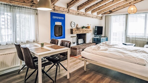 Moderni hotellihuone, jossa on näkyvillä puiset kattopalkit, ruokailutila pöytineen ja tuoleineen, sänky, televisio ja seinälle kiinnitetty kello.