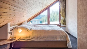 Ett sovrum med ett sluttande tak, en säng, ett skrivbord med en lampa och ett fönster med utsikt över träd och en byggnad.