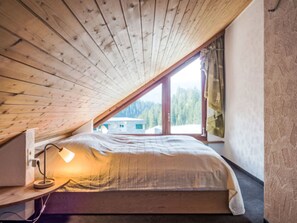 A bedroom with a sloped ceiling, a bed, a desk with a lamp, and a window with a view of trees and a building. - Val Signina 8-18 by Interhome (Laax)