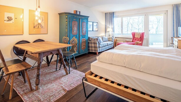 A room with a wooden bed, a blue cabinet, a plaid sofa, a dining table with chairs, and a window with curtains. - Quadra B223 by Interhome (Flims)