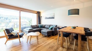 Uma sala de estar moderna com piso de madeira, sofá modular cinza, uma mesa de jantar com cadeiras e uma grande janela com vista para o exterior.