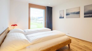 Uma cama de casal com estrutura de madeira, uma janela grande com vista para uma paisagem verde e três quadros pendurados na parede.