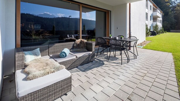 Uma área externa moderna com móveis de vime, uma porta de vidro que dá para uma vista de montanha e um pátio pavimentado.