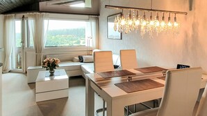 Ein modernes Zimmer mit Esstisch, Stühlen, einer Couch, einer Kristallleuchtre und Blick auf Grünflächen.