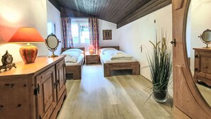Ein Zimmer mit zwei Betten, Holzboden, einem Fenster mit Vorhängen, einer Nachttischlampe, einem Spiegel, einem Schminktisch und einer Topfpflanze.