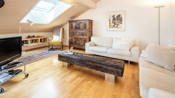 Een loft-achtige kamer met een schuine hemel, een houten plafond, een witte bank, een houten salontafel, een televisie op een standaard, een houten kast en een dakraam.