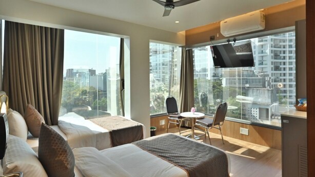 Quarto de hotel com duas camas, uma televisão, ventoinha no teto e janelas amplas com vista para a cidade.