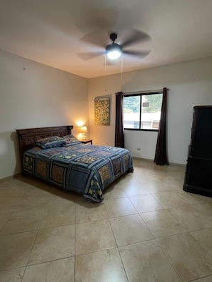 A bedroom with a bed, a ceiling fan, a window with curtains, and a nightstand with a lamp.
