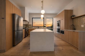 A modern kitchen with wooden cabinets, stainless steel appliances, a large island, and a view of the sunset.