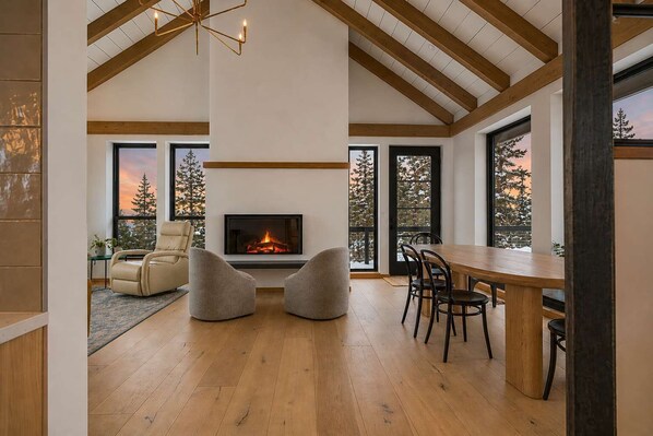 A spacious room with a fireplace, wooden flooring, a dining table with chairs, and large windows with a view of trees.