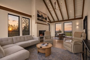 A spacious living room with a fireplace, large windows, and a high ceiling with exposed beams. - The Majestic - Elevated Views, Sauna & Game Room (Kalispell)