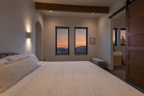 A bedroom with a large bed, two windows with a view, a bedside lamp, and a wooden ceiling beam.