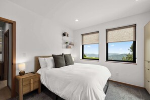 Une chambre à coucher avec un lit, une table de chevet et deux fenêtres donnant sur un beau paysage.