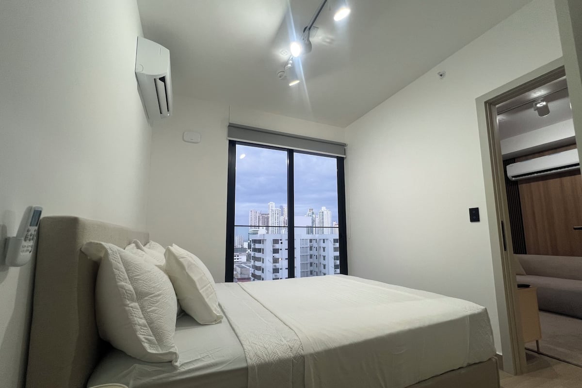 Un dormitorio con cama, ventana con vista a la ciudad, unidad de aire acondicionado y puerta que da a otra habitación.