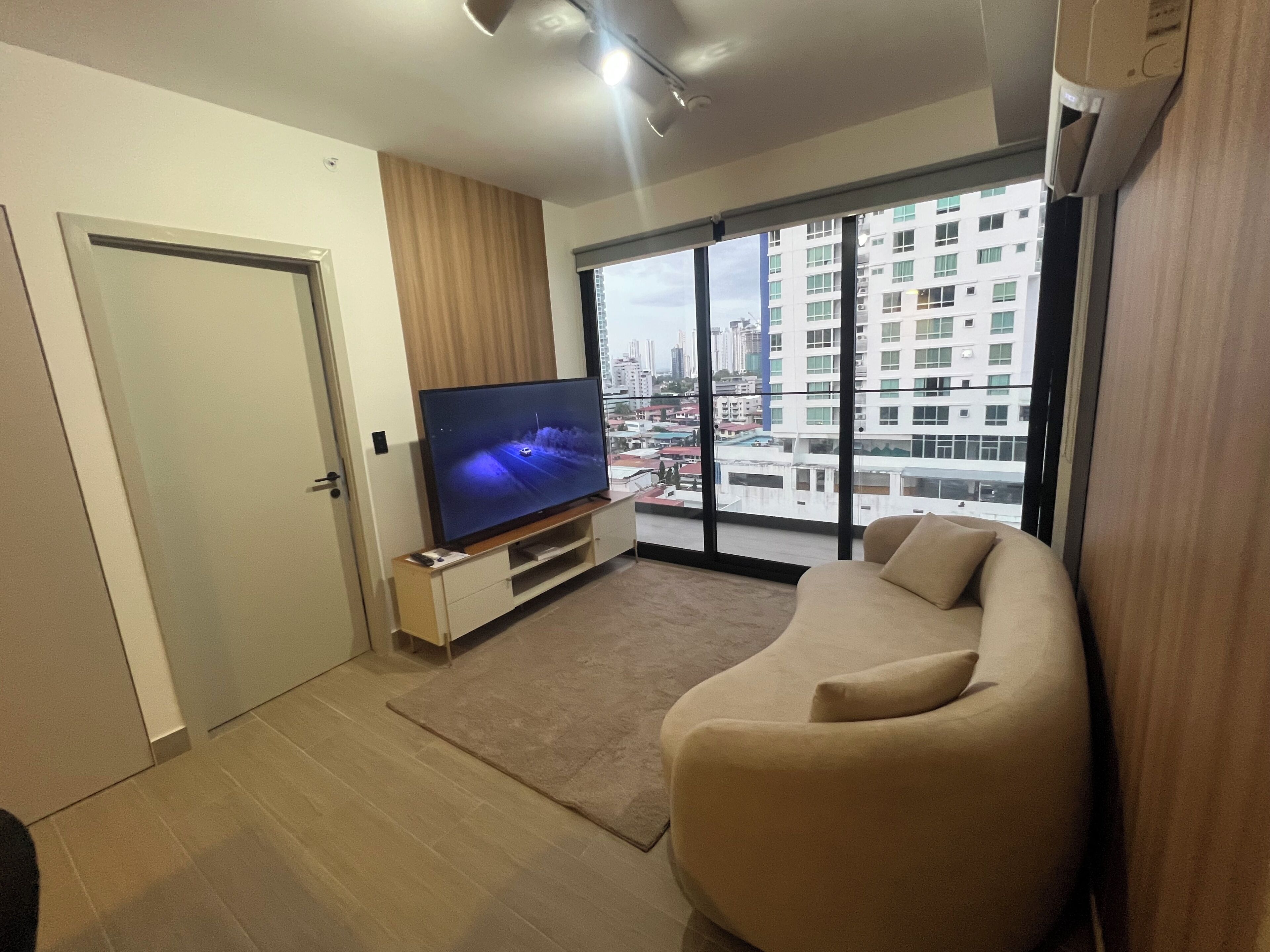 A modern living room with a flat-screen TV, a beige sofa, a large window with a city view, and a door.
