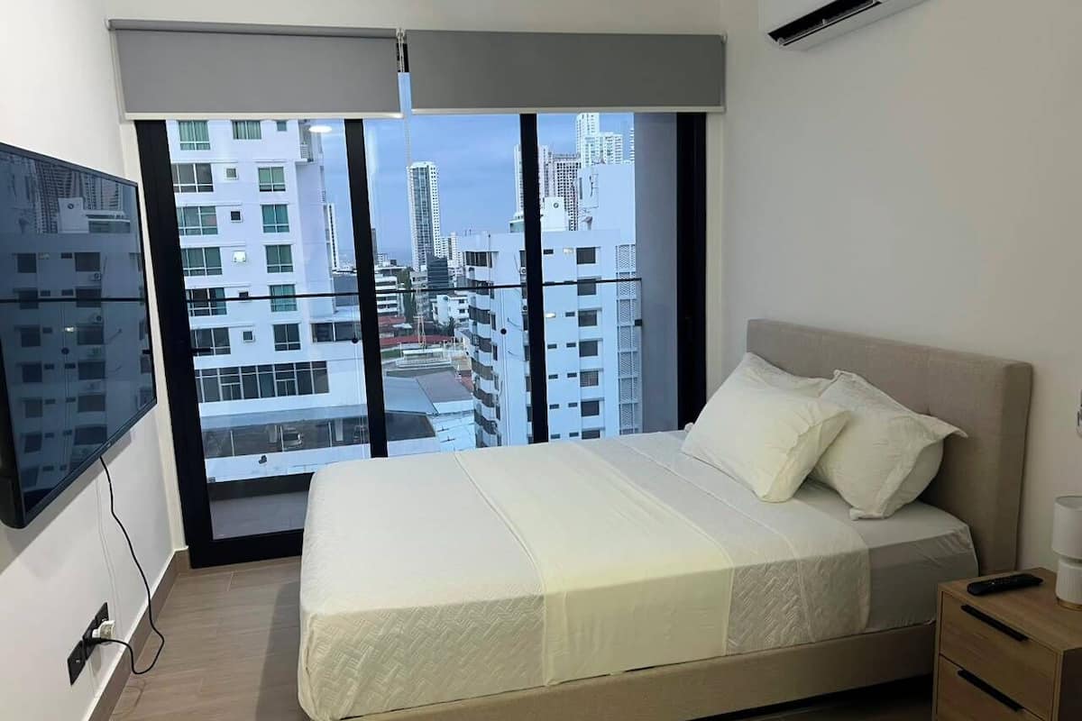 Un dormitorio con cama, televisor, mesita de noche y ventana con vistas a la ciudad.
