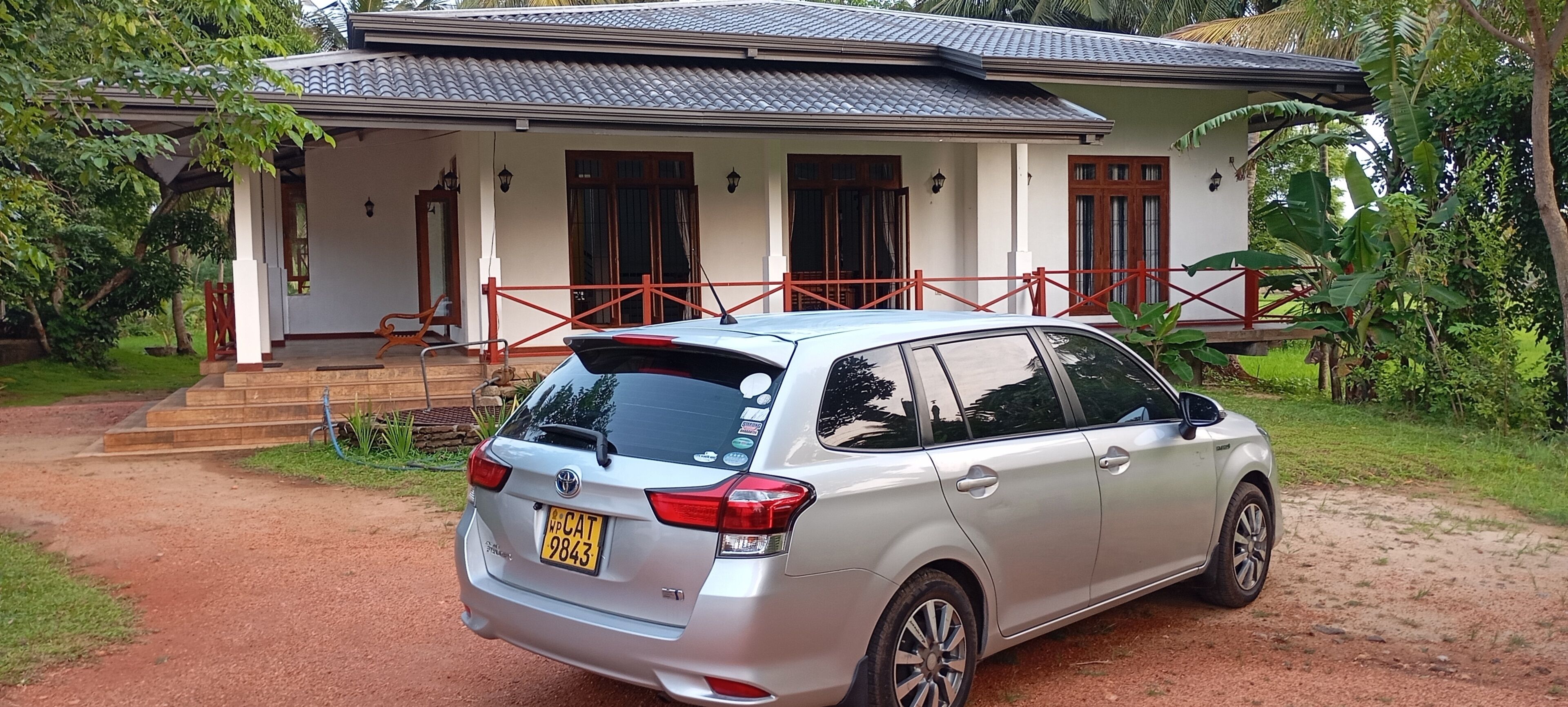 A silver car parked in front of a house with a red fence, a porch, and a garden.