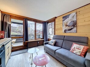 Ein gemütliches Zimmer mit grauem Sofa, einem kleinen Tisch, einem Heizkörper und einem Fenster mit Vorhängen.