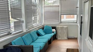 A sunroom with a long, dark-colored sofa, a ceiling fan, and large windows with blinds.