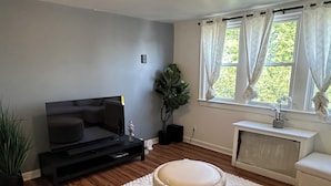 Een woonkamer met een flatscreen televisie, een rond poefje, een potplant en een raam met gordijnen.