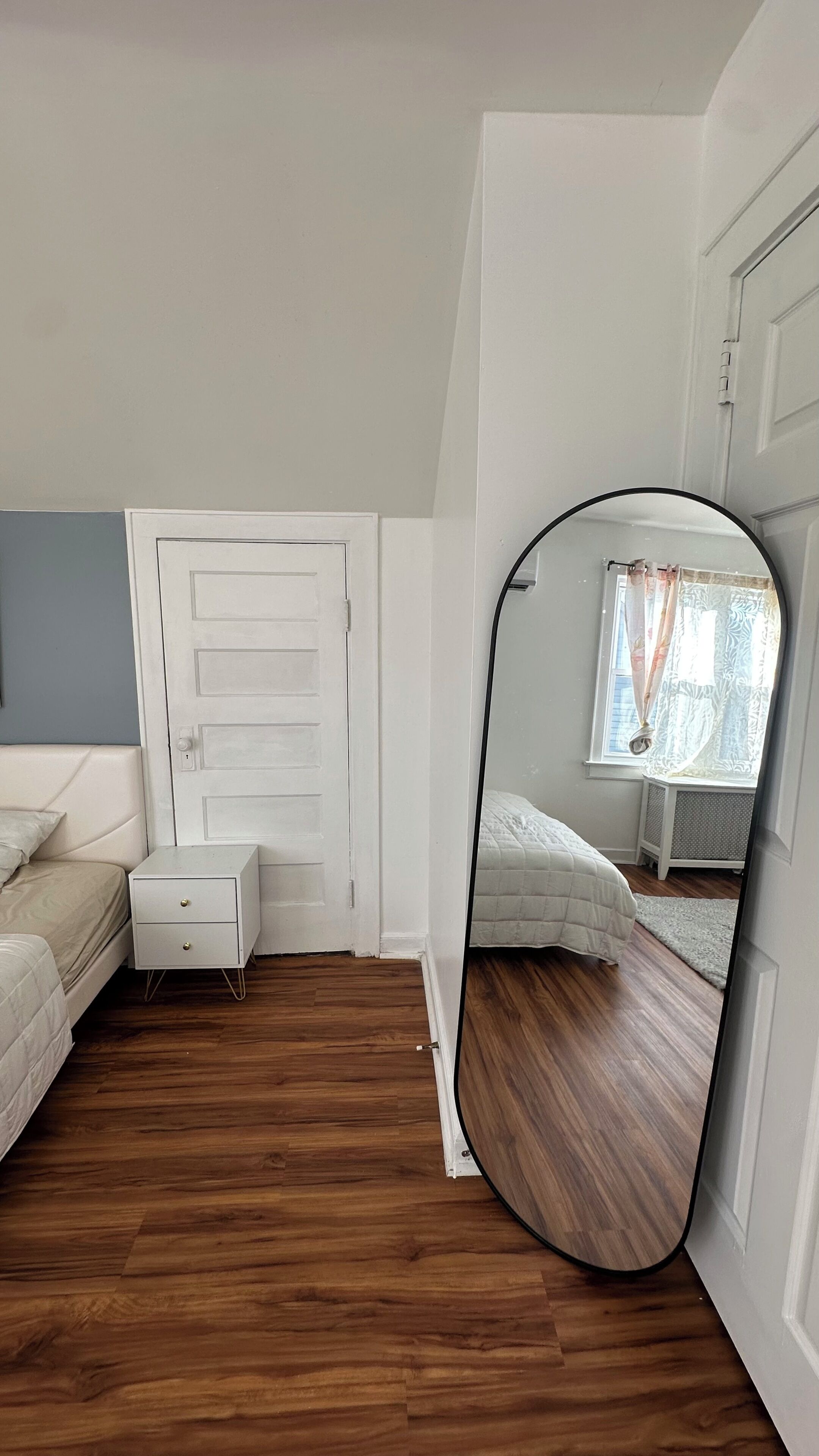 A bedroom with a wooden floor, a white door, a white bedside table with drawers, a bed with a white duvet, and a large mirror reflecting the room.