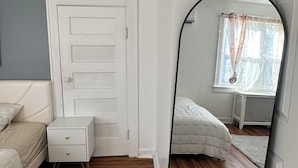 Een slaapkamer met houten vloer, een witte deur, een witte nachtkastje met lades, een bed met witte dekbedovertrek, en een grote spiegel die de kamer weerspiegelt.