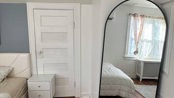 A bedroom with a wooden floor, a white door, a white bedside table with drawers, a bed with a white duvet, and a large mirror reflecting the room.