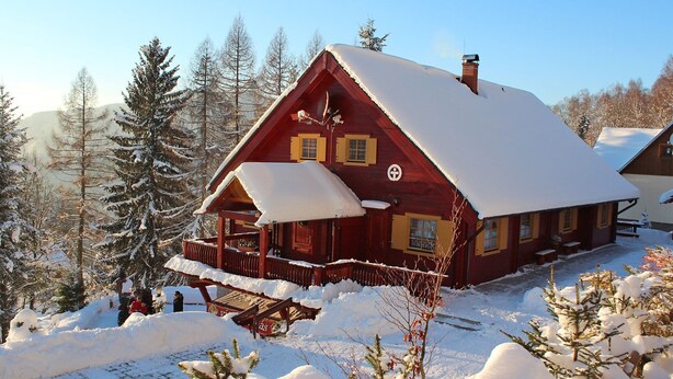 Eine rote Holzhütte mit schneebedecktem Dach, umgeben von mit Schnee beladenen Bäumen und blauem Himmel.