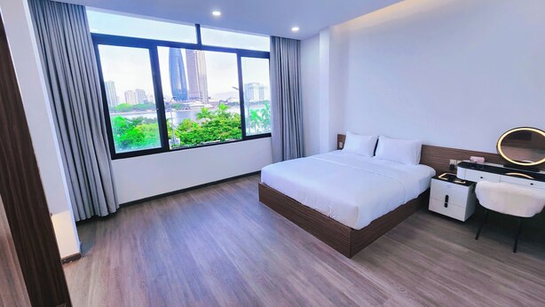 A modern hotel room with a large bed, a bedside table with a mirror, and a view of a cityscape through large windows. - Min Sweet Home 2 (Da Nang)