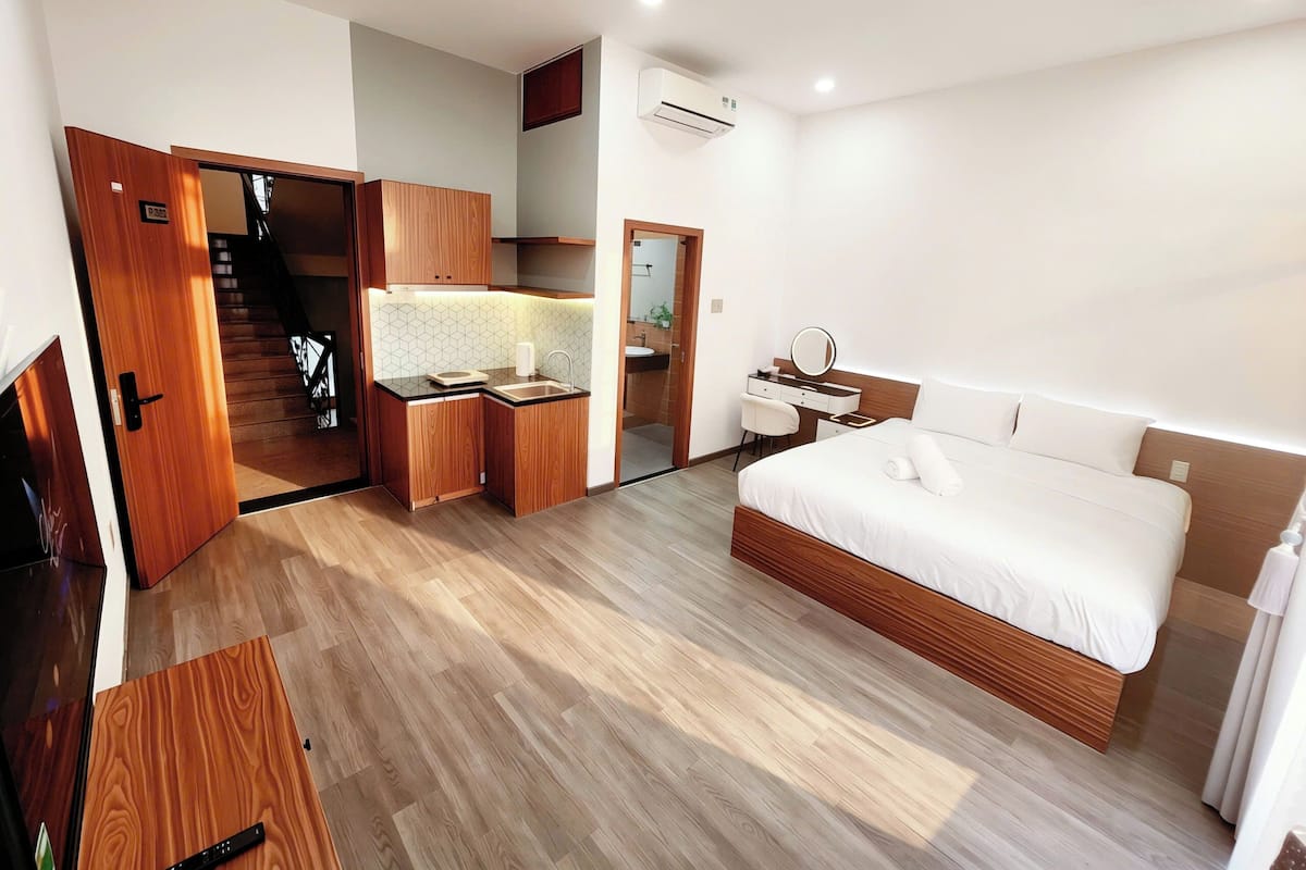 Una habitación de hotel moderna con suelo de madera, una cama, un escritorio con espejo, una kitchenette pequeña y un baño.