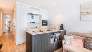 Een moderne keuken met een inbouwoven, een koffiezetapparaat en een spoelbak. Een boekenkast met boeken en decoratieve objecten is zichtbaar. In de voorgrond staat een bank met kussens.