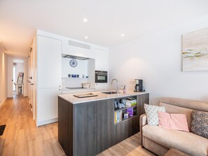 Eine moderne Küche mit Einbauofen, Kaffeemaschine und Spüle. Ein Bücherregal mit Büchern und Dekorationselementen ist zu sehen. Im Vordergrund steht ein Sofa mit Kissen.