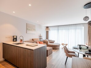 Eine moderne Küche mit Kochinsel, ein Essbereich mit rundem Tisch und ein Wohnbereich mit Sofa und Hocker.