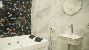 Une salle de bain avec une baignoire blanche, un lavabo rectangulaire orné de motifs dorés, un miroir et un mur recouvert de carreaux aux motifs géométriques.