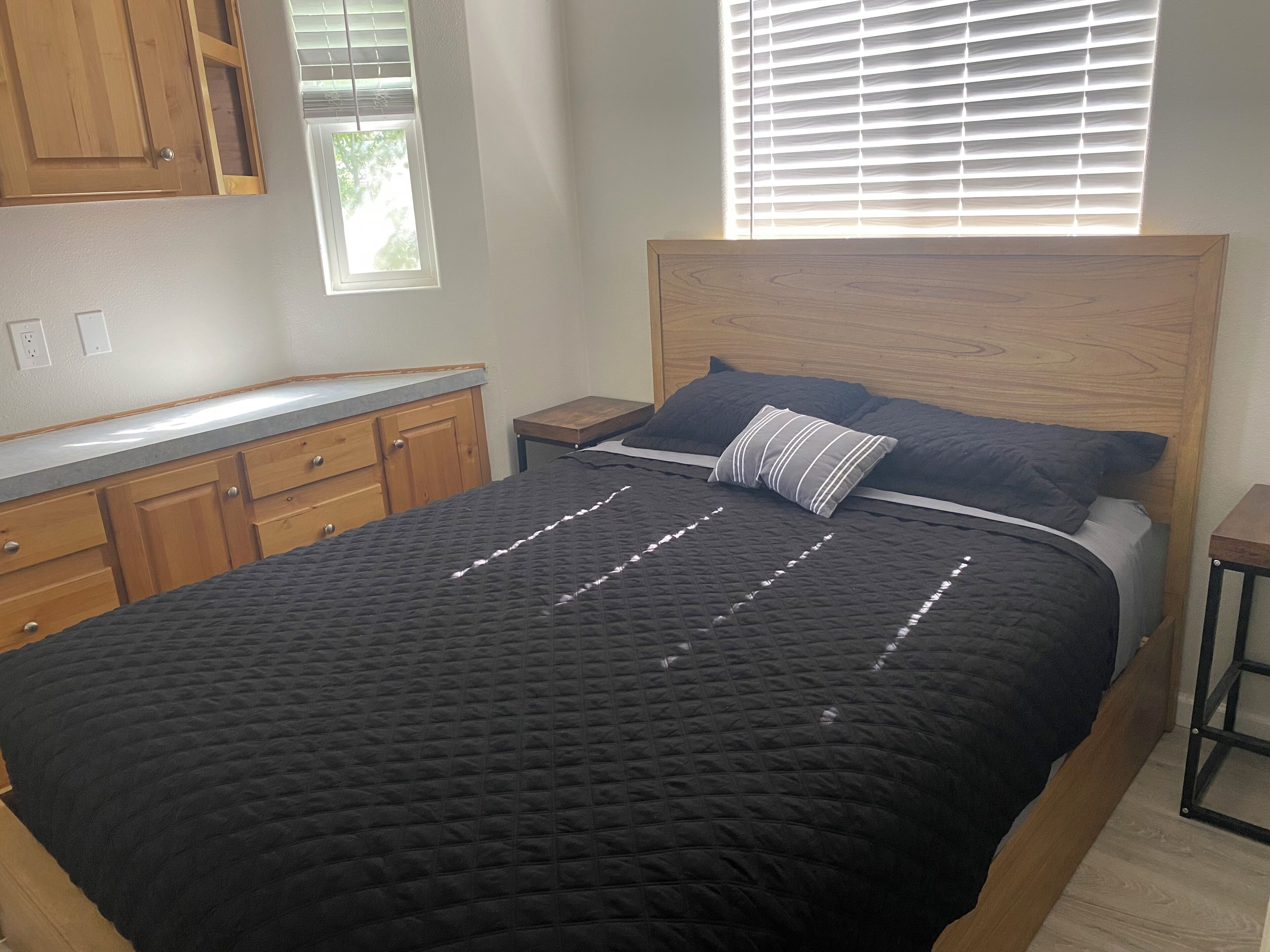 A bedroom with a bed, wooden headboard, a nightstand, a window with blinds, and a wooden dresser.