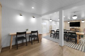 A spacious room with a wooden dining table, black chairs, a TV mounted on the wall, and a large window.