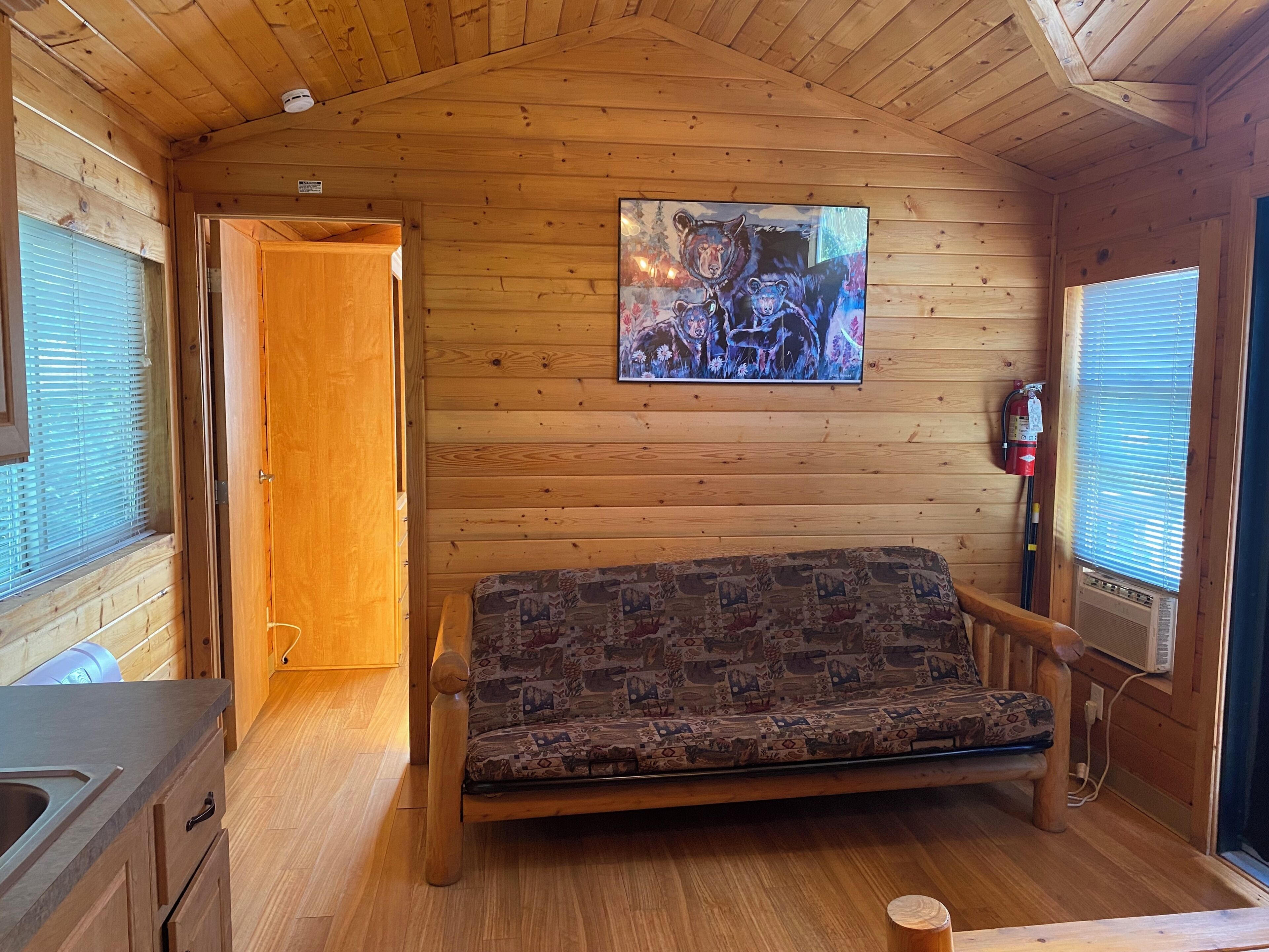 Interior de una cabaña de madera con un sofá, un extintor y una ventana con persianas.