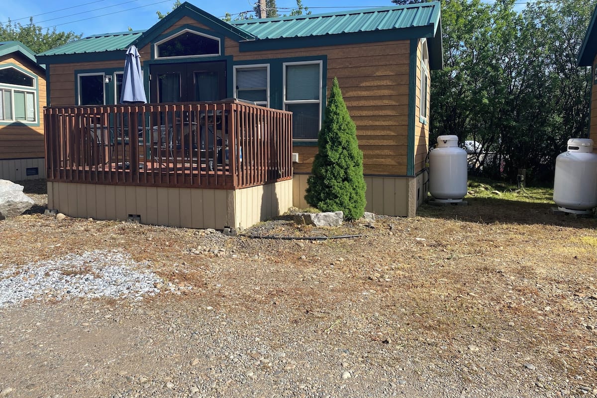 A small wooden cabin with a fenced deck, a tall coniferous tree, and two large propane tanks.