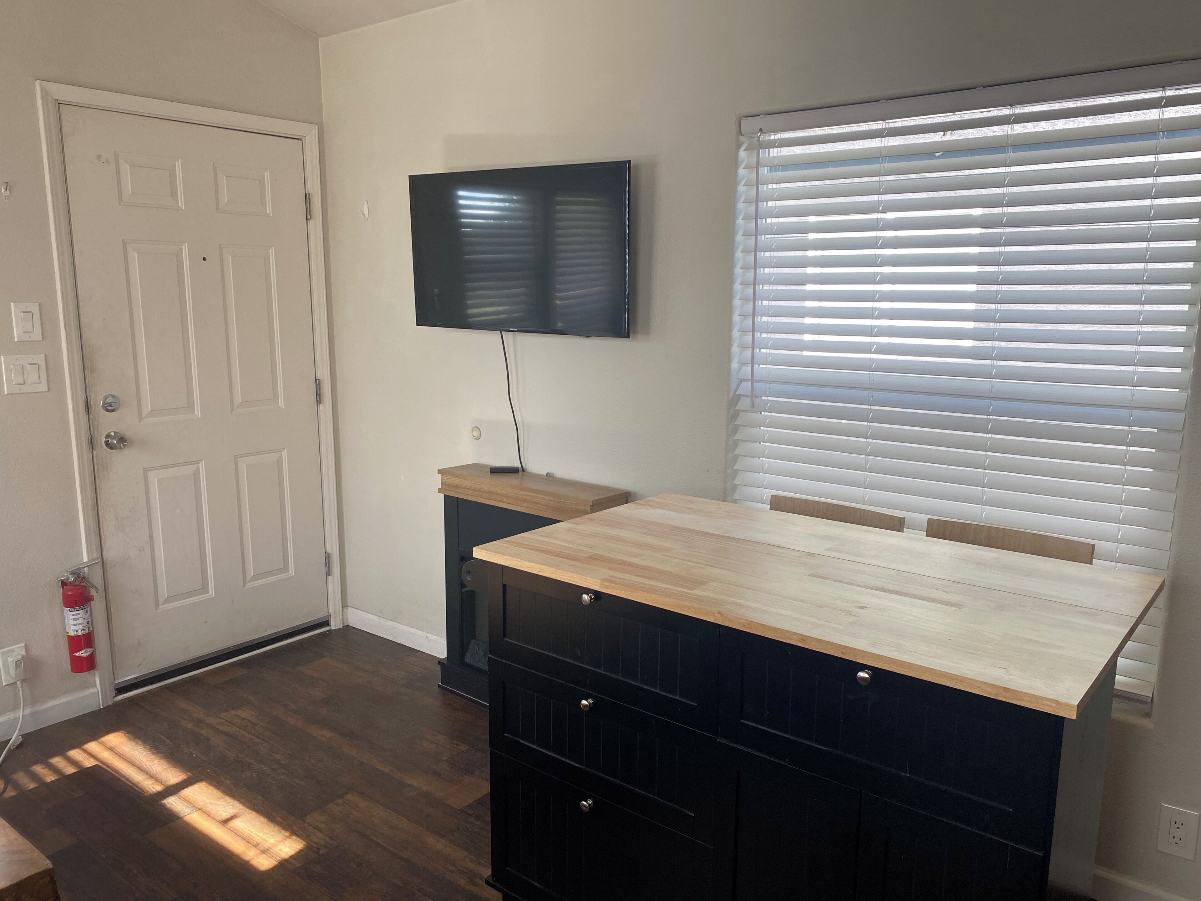 A room with a wooden desk, a television mounted on the wall, a door, and a window with blinds.