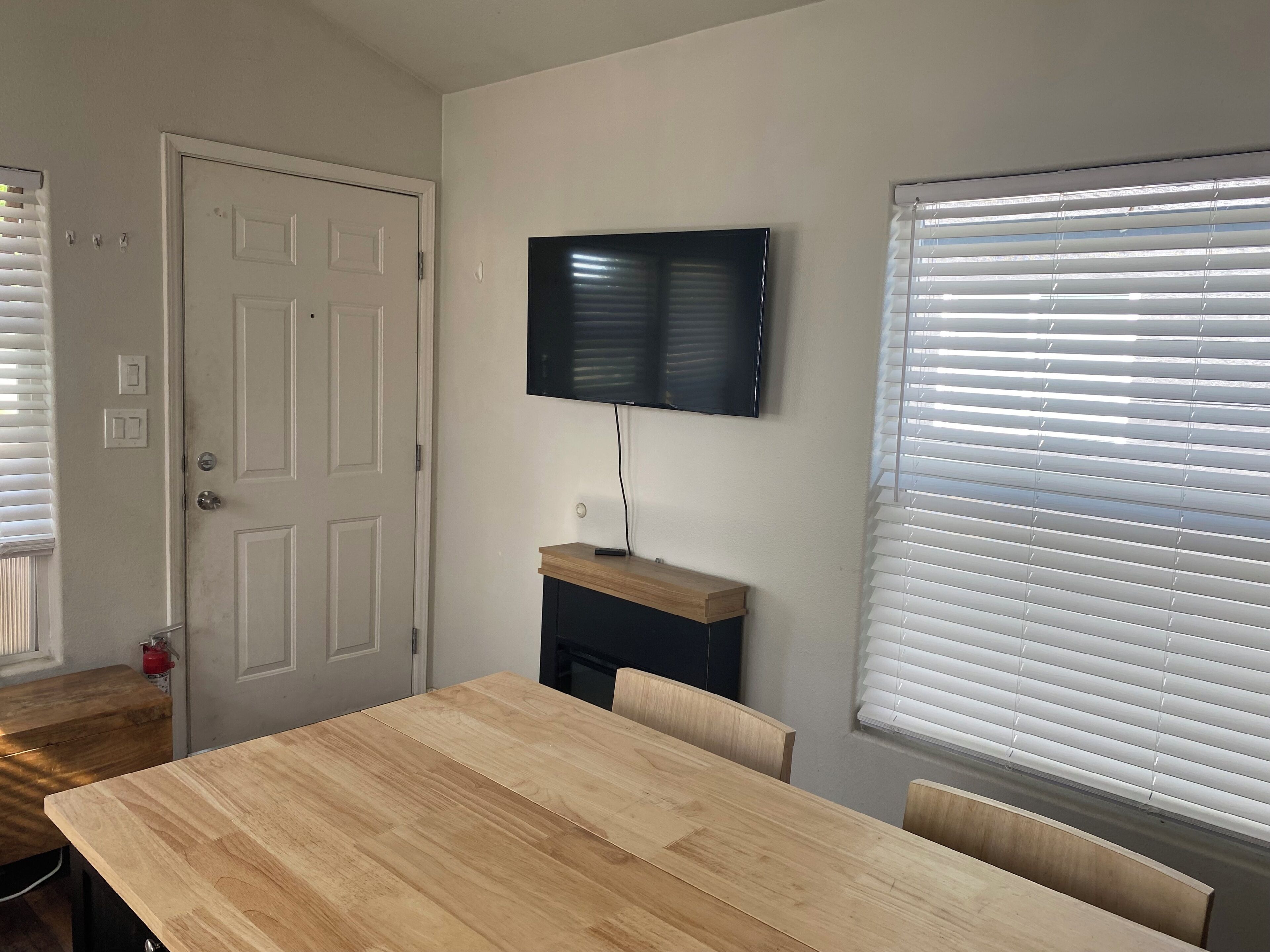 Una habitación con una mesa de comedor de madera, un televisor montado en la pared y una ventana con persianas.