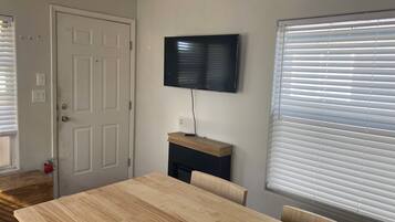 Una habitación con una mesa de comedor de madera, un televisor montado en la pared y una ventana con persianas.