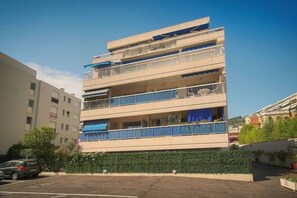 A multi-story residential building with balconies, a parking area, and a green hedge in front. - Frgk Alexander III Elegant 1-Bedroom Apartment with Terrace in Palm Beach (Cannes)