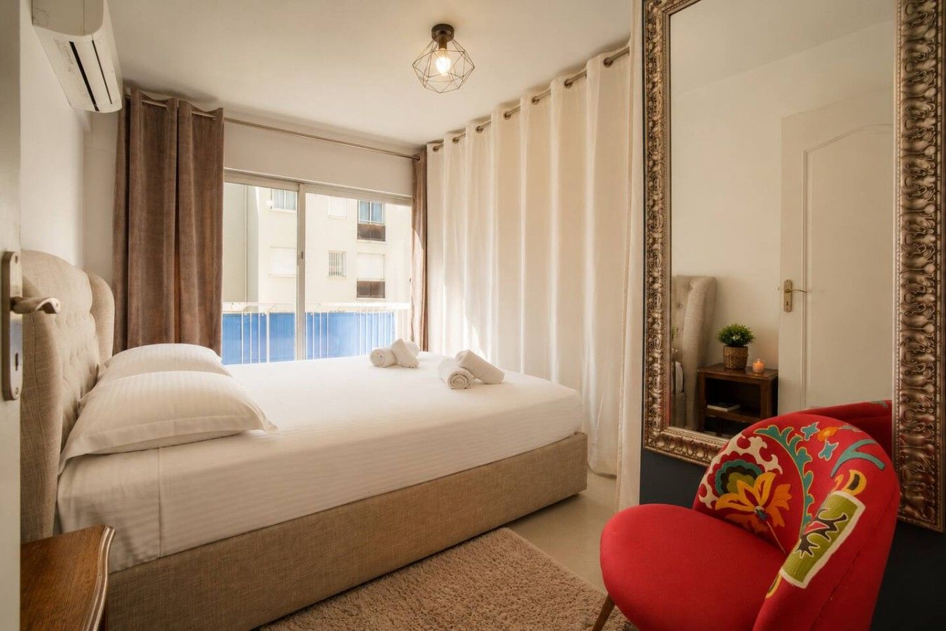 A hotel room with a bed, a red chair, a large mirror, and a window with curtains.